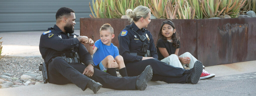 Two PHX police officers sitting on the ground talking with children outside.