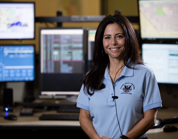 A communications operator woman in a blue shirt standing in front of several monitors.