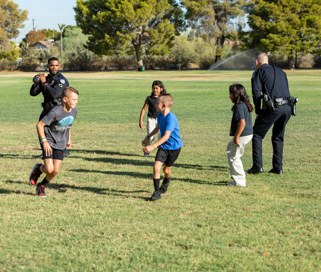 A group of kids playing a game of football in a park with two PHX PD police officers.